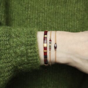 Een pols met drie armbanden. Links een armband met platte glaskralen in roodtinten met goud, in het midden een gouden schakelarmband met een mix aan edelsteentjes en rechts een gouden schakelarmband met rood granaat ruitje.