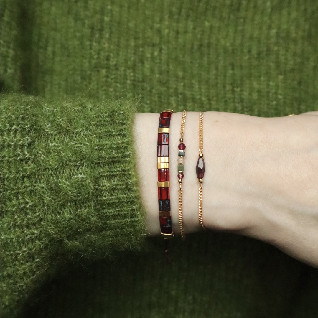 Een pols met drie armbanden. Links een armband met platte glaskralen in roodtinten met goud, in het midden een gouden schakelarmband met een mix aan edelsteentjes en rechts een gouden schakelarmband met rood granaat ruitje.