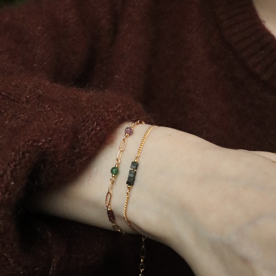 Een pols met twee goudkleurige armbanden. Schakelarmband Nani met een mix van groene en roze edelstenen en Linden met schijfjes groene zandsteen.